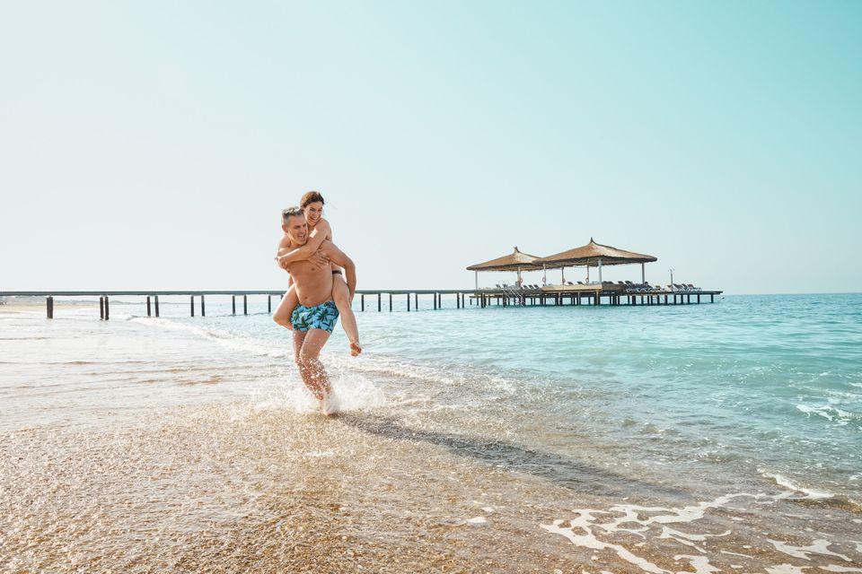 Das Bild zeigt ein Paar am Strand auf den Malediven mit einem Steg im Hintergrund, dabei trägt der Mann seine Partnerin Huckepack.